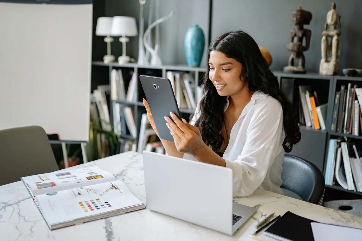 A woman using a digital tablet beside her laptop at a work desk.
