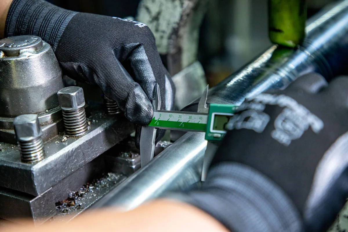 Gloved hands measuring a metal component with a vernier caliper at a machine.