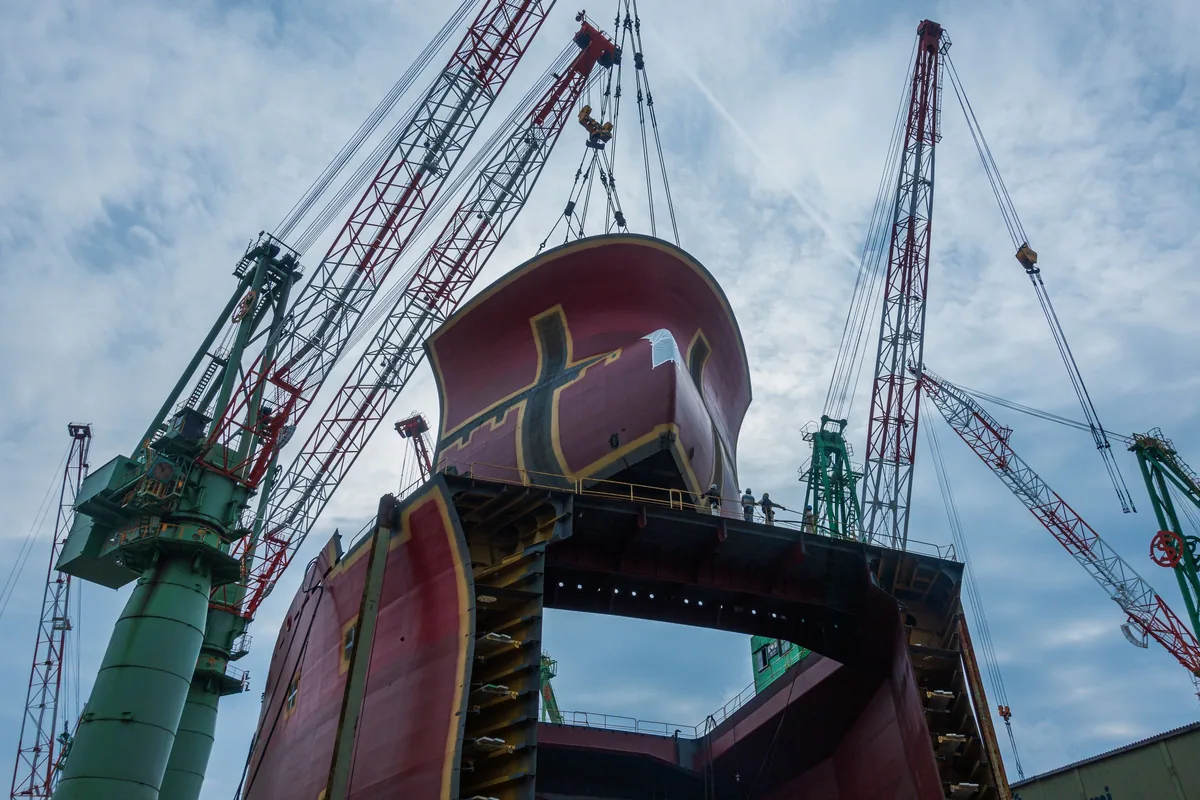 Cranes lifting a large red hull section at a shipyard.